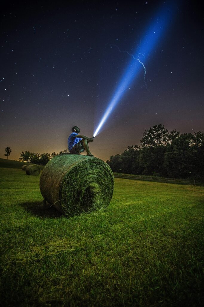 A man sits on a hay bale in a field, shining a flashlight into the night sky full of stars.