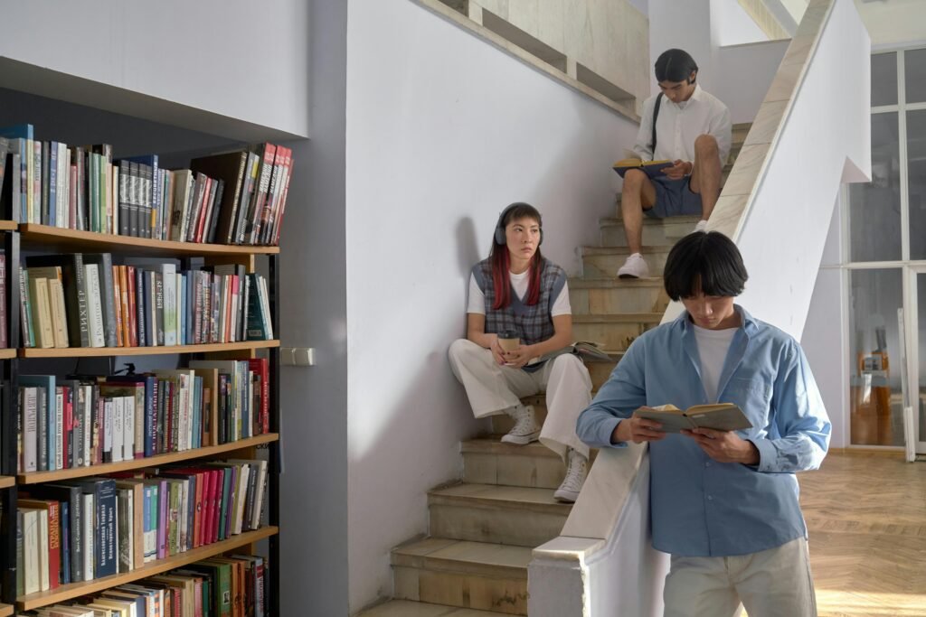 Three students reading on a staircase near bookshelves inside a library setting.